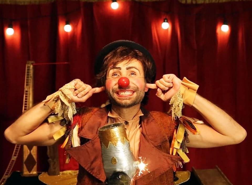 Há um homem sorrindo, com dedos no ouvido e caracterizado de palhaço, usando o tradicional nariz vermelho e um chapéu preto. Atrás dele, há uma cortina vermelha típica de teatro de picadeiro e luzes pendurados. 