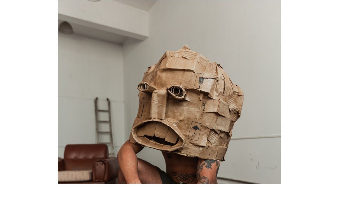 Há um homem agachado no chão, apoiado sobre um dos pés, usando um tênis verde, uma bermuda verde e está sem camiseta. Ele possui tatuagens pelo braço. Não se vê o seu rosto, que está encoberto por uma máscara feita de um material similar ao papelão. A feição da máscara possui dois olhos, um nariz reto e uma boca aberta com dentes. Atrás dele, há um sofá marrom e uma escada de obra. Ele está sobre um piso de tablado e cercado de paredes brancas.