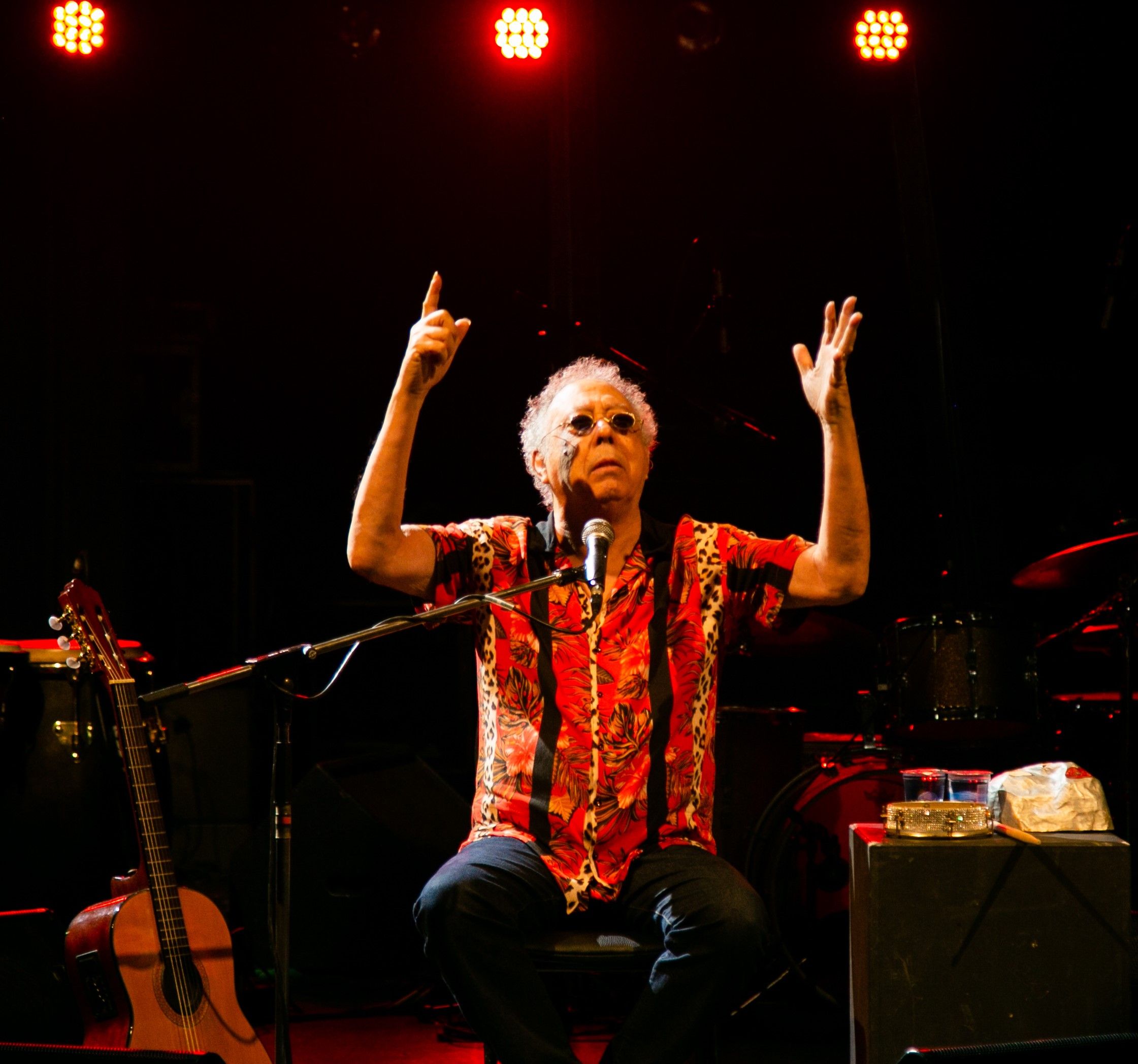 Foto colorida de Jards Macalé no palco. O músico, um homem branco de cabelo grisalho cacheado e óculos escuros, está sentado de frente, vestindo uma camisa de manga curta com estampa vermelha e preta. Ele está em frente a um microfone, com o braço direito levantado e o dedo indicador apontado para cima, e a mão esquerda também erguida, com a palma aberta. O fundo está escuro, com luzes de palco vermelhas brilhando acima. À sua esquerda, vê-se parte de um violão e, à direita, uma pequena mesa com um pandeiro.