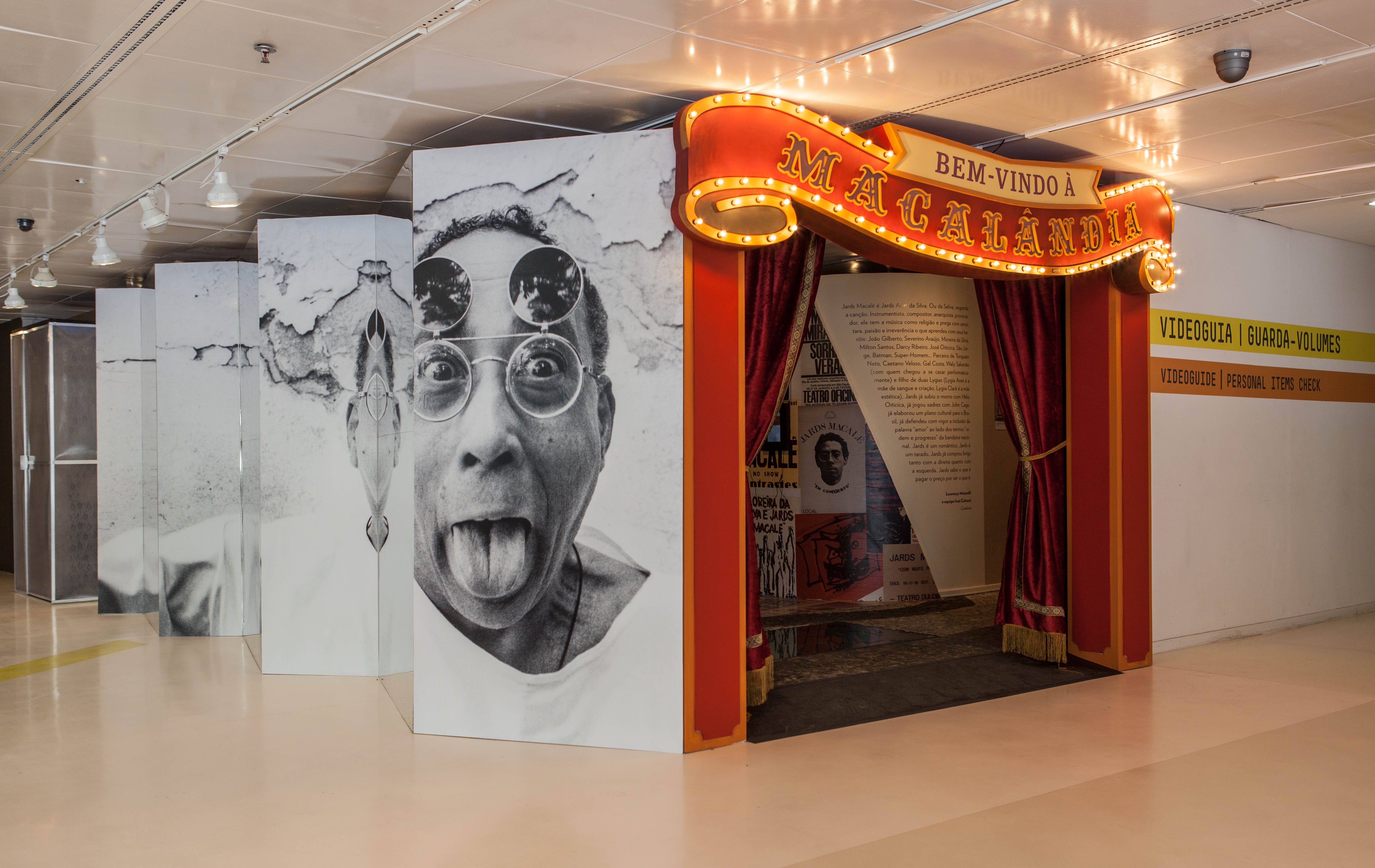 Foto colorida da entrada da exposição Ocupação Jards Macalé. À esquerda, um grande painel sanfonado exibe uma foto em preto e branco de Jards Macalé fazendo careta, com a língua para fora e usando óculos redondos. À direita, um portal de entrada estilo circo, com um letreiro luminoso em arco onde se lê 