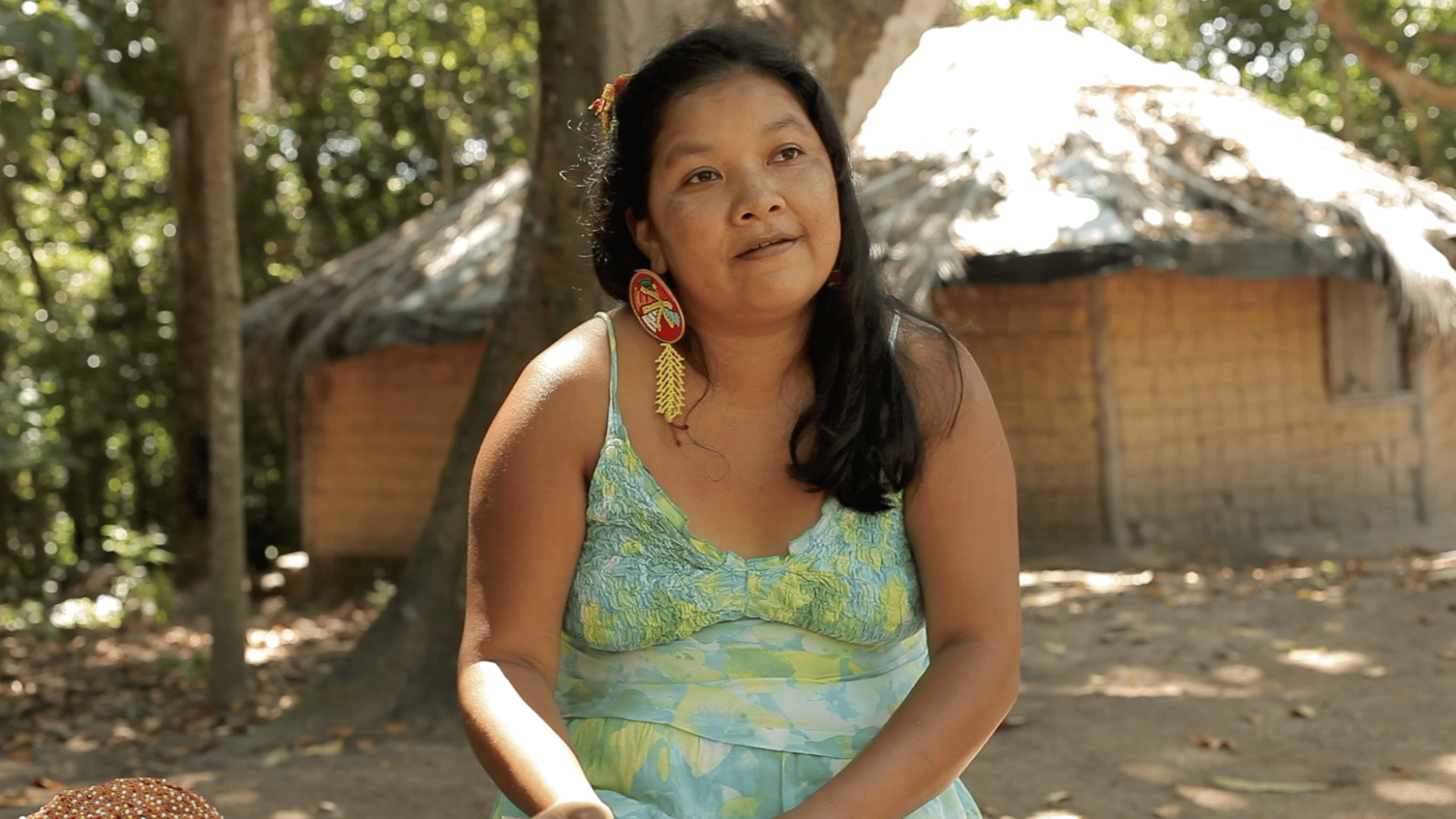 Sueli Maxacali est&aacute; sentada. Ela usa um vestido de alcinha azul claro, com detalhes em verde, amarelo e branco. Ao fundo, h&aacute; &aacute;vores.