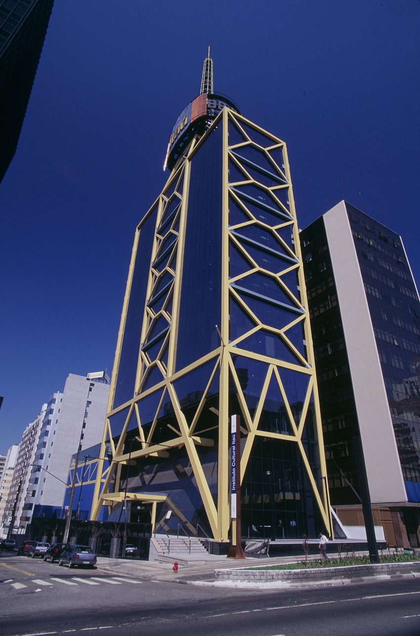A foto mostra um prédio alto e moderno, com fachada de vidro azul escuro e uma estrutura externa amarela formando padrões geométricos triangulares. No topo há um cilindro com letreiros e uma antena. O edifício se destaca entre construções vizinhas mais simples. O céu está claro e azul, e carros e pedestres aparecem na rua em frente ao prédio.