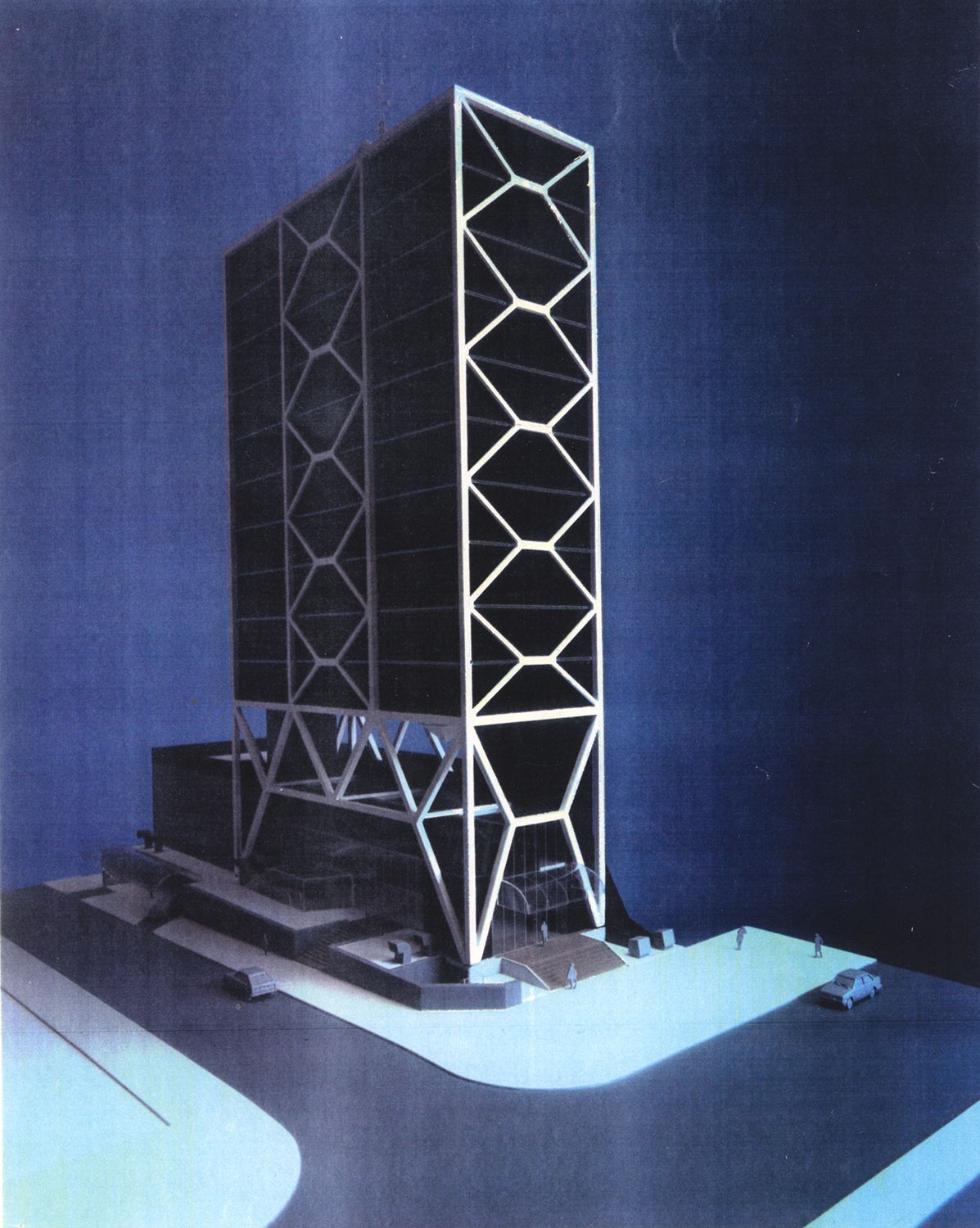 A imagem mostra uma maquete arquitetônica de um prédio alto e moderno. A torre tem fachada escura, envolvida por uma estrutura externa branca em formas geométricas repetidas. A base do edifício é mais larga e possui rampas e escadas. Há pequenas figuras humanas e um carro, ajudando a indicar a escala. O fundo é azul e uniforme, destacando a construção.