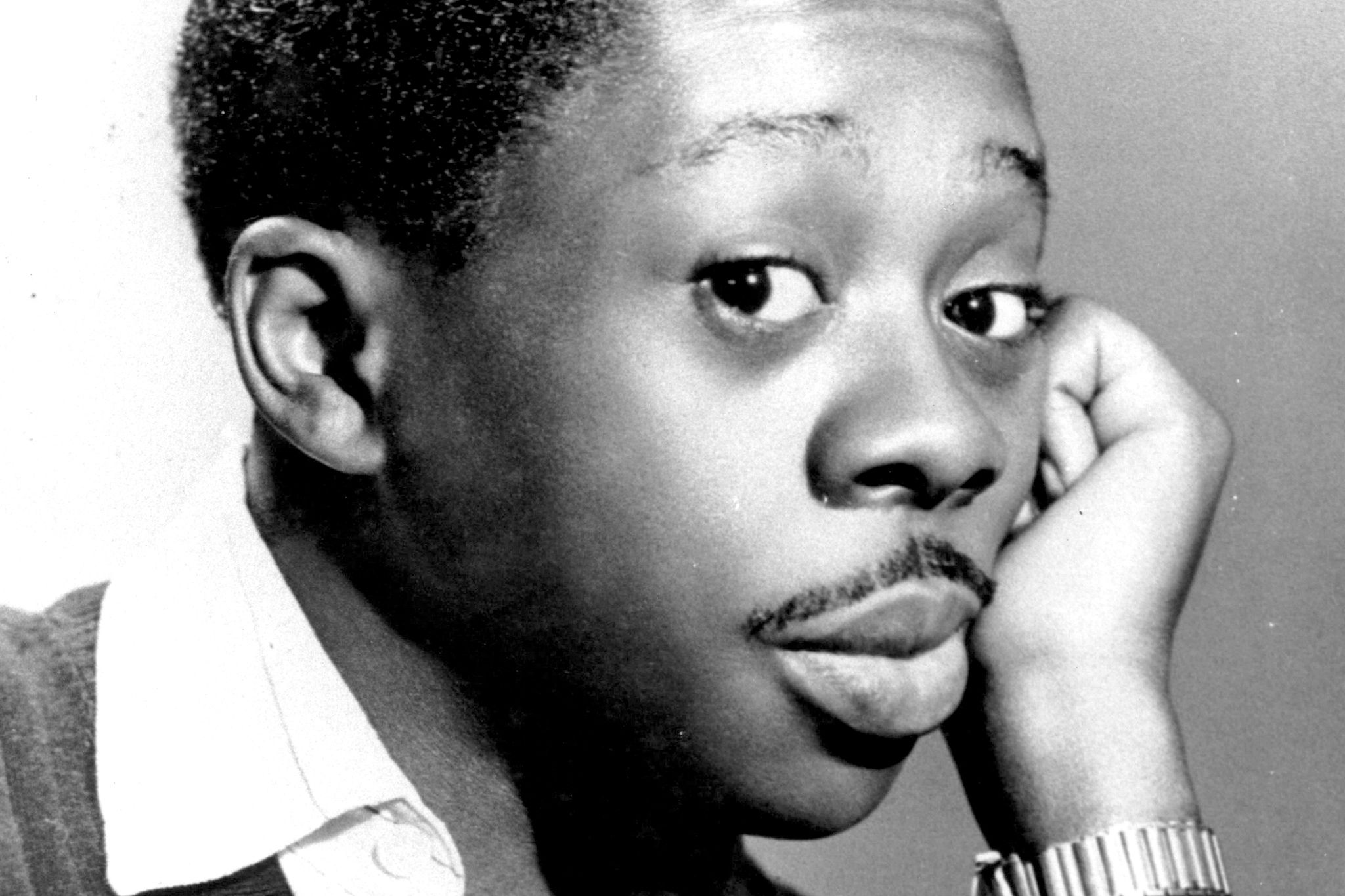 A foto é em preto e branco e mostra Grande Othelo jovem, com expressão pensativa. Ele apoia o rosto na mão, inclinando ligeiramente a cabeça, e traz um olhar direcionado para a câmera. O artista veste uma camisa com gola. No pulso esquerdo, usa um relógio. O fundo é neutro.