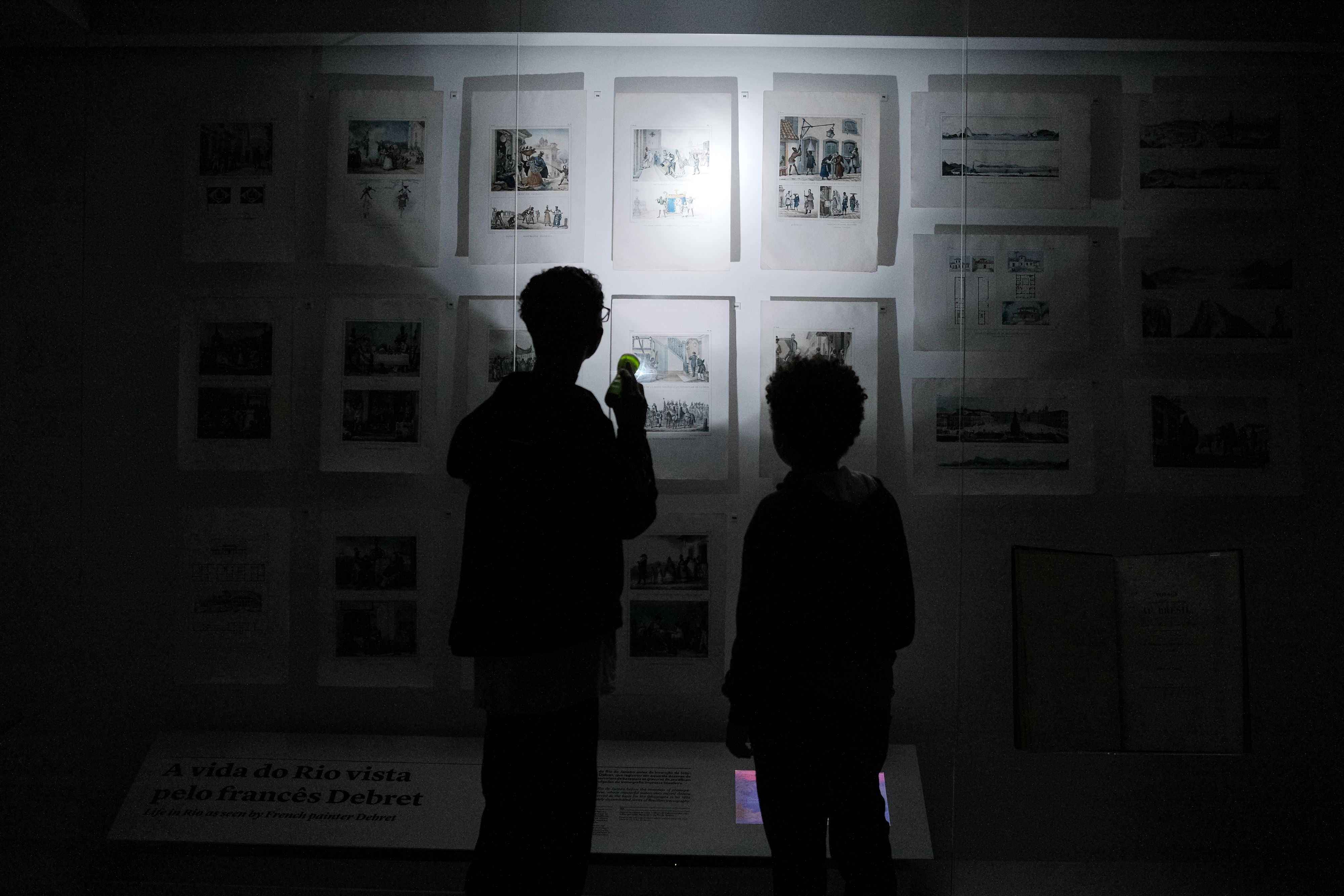 A foto mostra duas pessoas vistas de costas, em um ambiente escuro de exposição. Elas observam uma parede com várias gravuras iluminadas suavemente por uma lanterna. A pessoa à esquerda segura uma pequena lanterna verde que destaca uma das imagens. As silhuetas aparecem em primeiro plano, enquanto os desenhos expostos — cenas históricas do Rio de Janeiro — ocupam toda a parede à frente. Uma placa no canto inferior menciona o pintor francês Debret. A foto mostra duas pessoas vistas de costas, em um ambiente escuro de exposição. Elas observam uma parede com várias gravuras iluminadas suavemente por uma lanterna. A pessoa à esquerda segura uma pequena lanterna verde que destaca uma das imagens. As silhuetas aparecem em primeiro plano, enquanto os desenhos expostos — cenas históricas do Rio de Janeiro — ocupam toda a parede à frente. Uma placa no canto inferior menciona o pintor francês Debret.