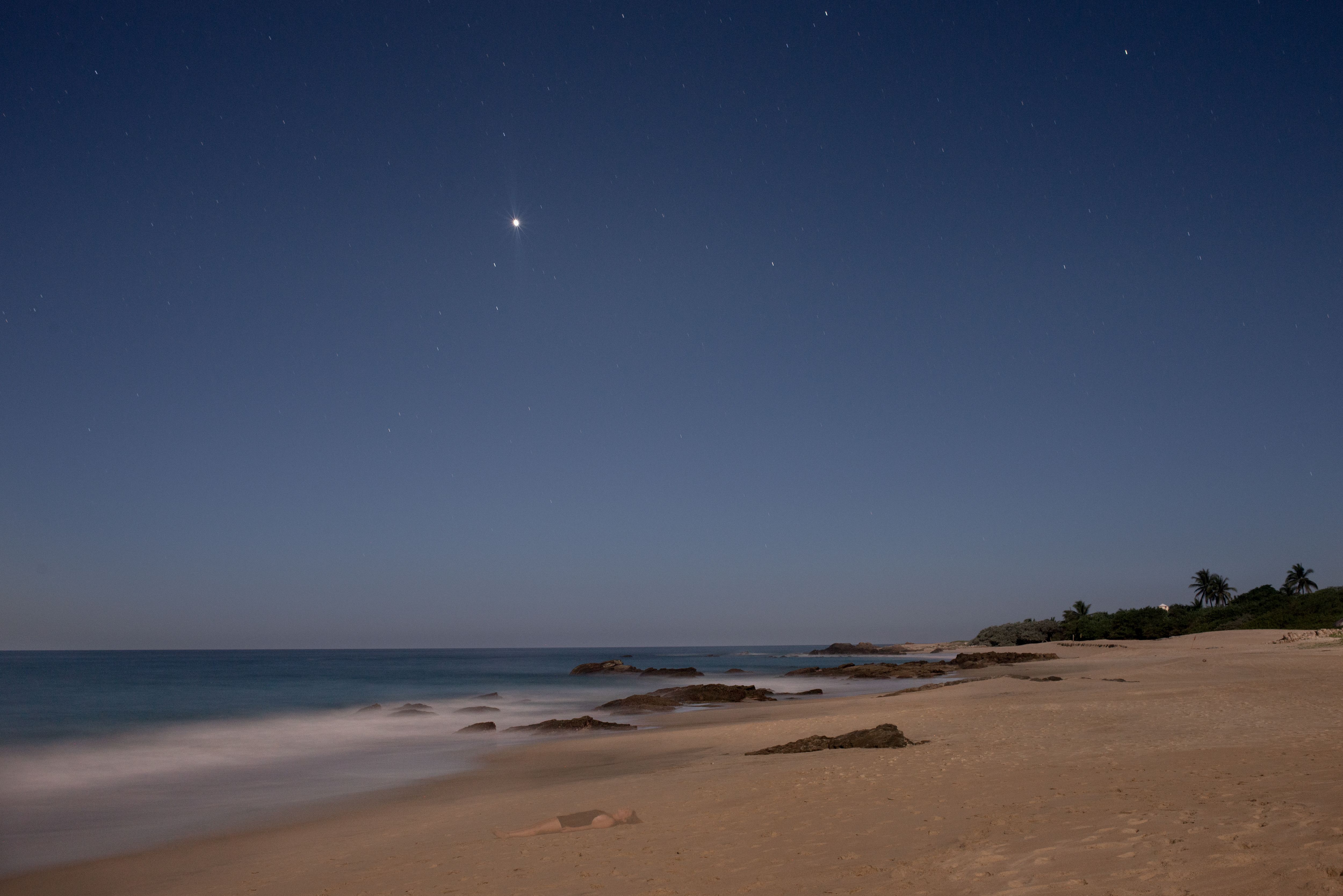 Faixa de areia e mar, com algumas pedras. Na areia tem um corpo deitado e desfocado. Mais ao fundo o céu ao entardecer com algumas estrelas.