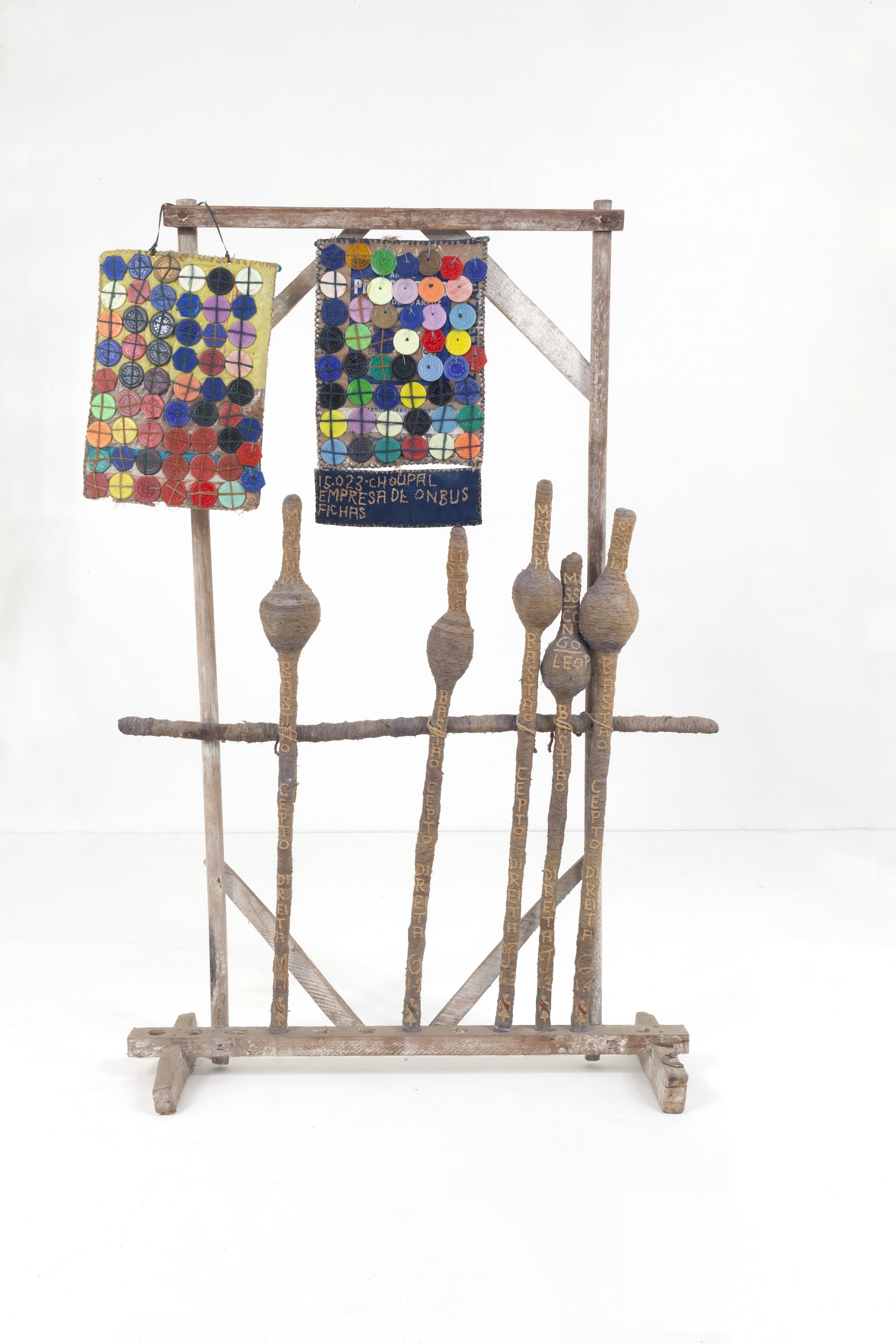 Obra do artista Bispo do Rosário, chamada [Recordações]. A obra é feita em madeira, em formato retangular, ela está em pé. Dentro dela existem dois pedados de tecidos também retangulares, com botões coloridos pregados nele. Encostado na obra, existem diversas cajados encostados. 